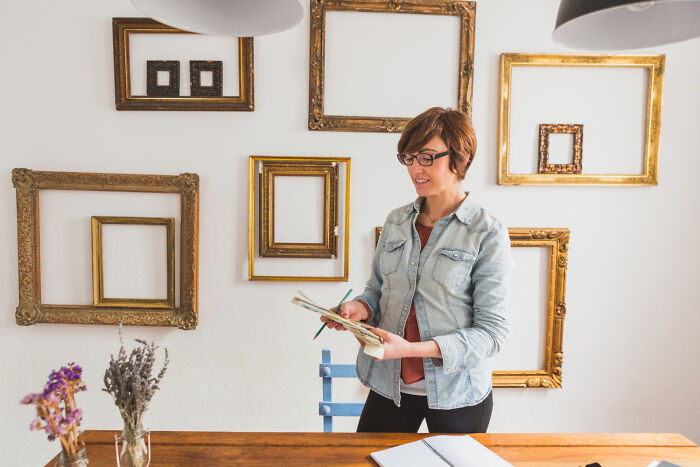 Woman in glasses standing by a table with empty picture frames on the wall, representing brutal wake-up calls.