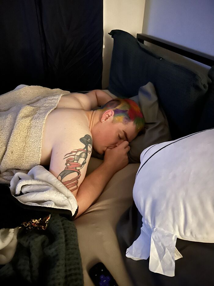 Man lying shirtless on bed with colorful hair and tattoo, showcasing awkward body language in a casual setting.