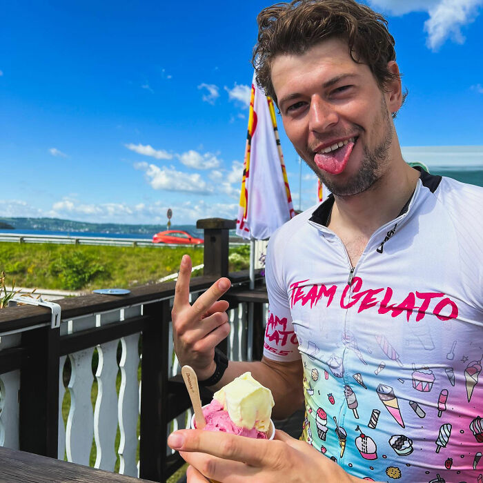 Young man in a Team Gelato cycling jersey holding ice cream, outdoors near a waterfront, expressing playful mood.