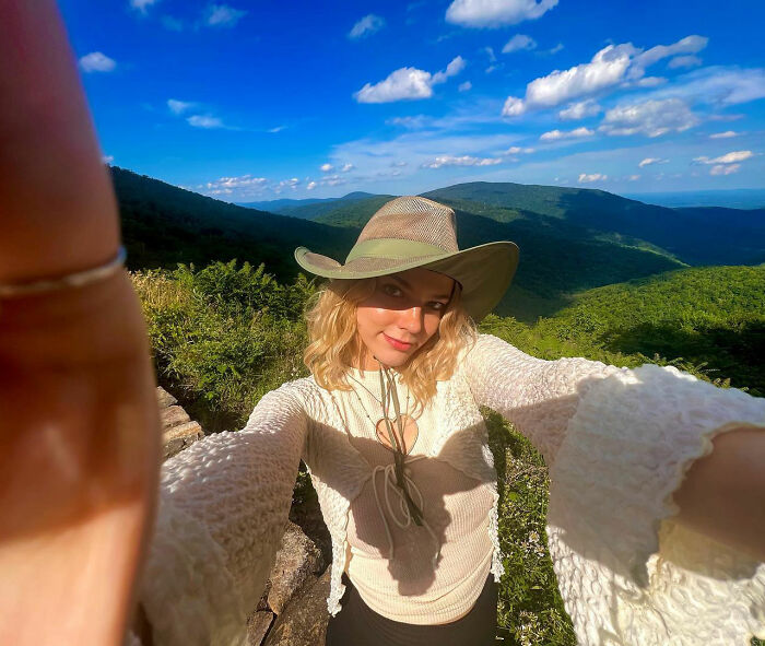 Young woman hiking New York's highest peak wearing a hat, taking a selfie with mountainous landscape and blue sky.