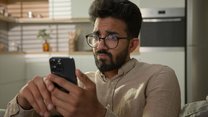 Man with glasses in a beige shirt looking skeptically at his phone, reflecting wildest dating experiences and reactions.