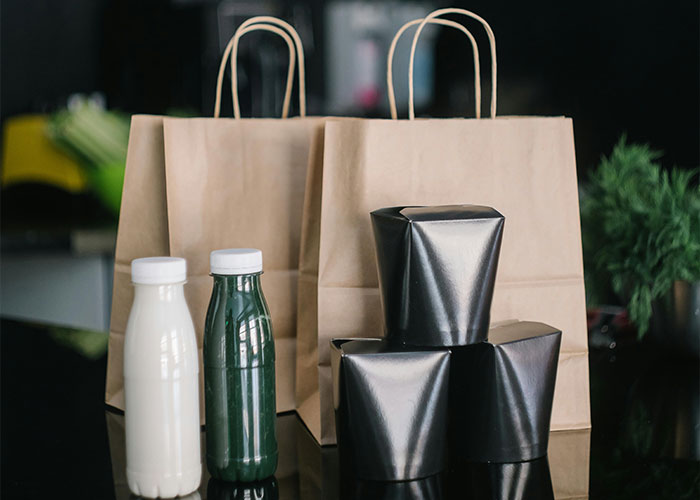Paper lunch bags with bottled drinks arranged on a black surface representing coworker lunches and entitlement conflict.