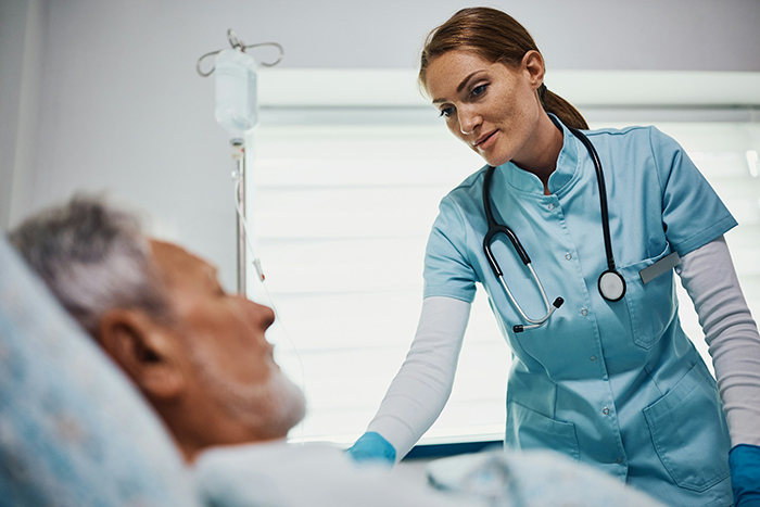 Female psychiatric nurse in blue scrubs caring for an elderly male patient in a hospital room setting.