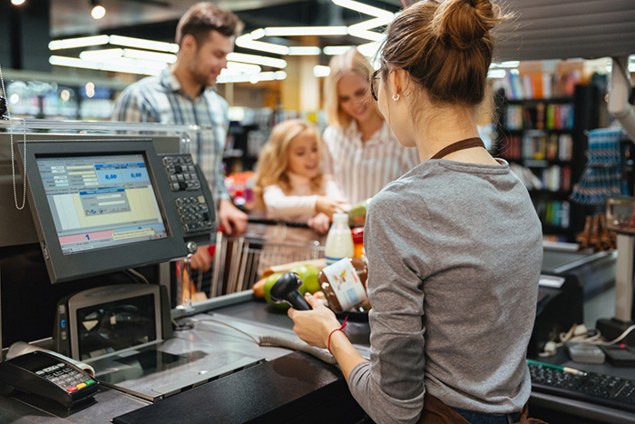 Cashier scanning groceries at checkout counter while customers wait during a busy store shift crisis.