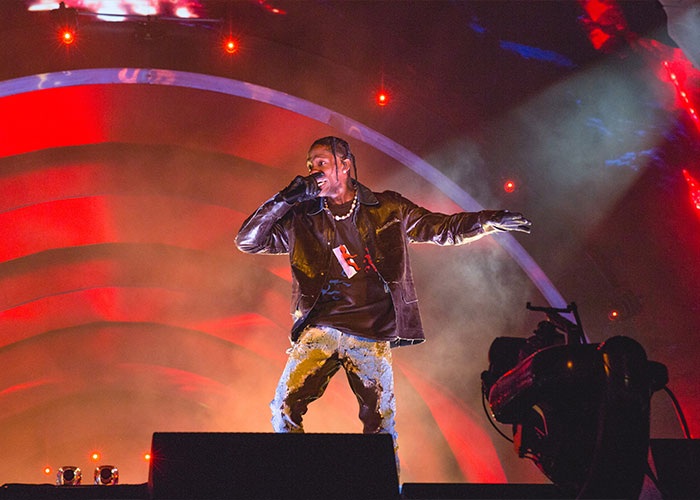 Travis Scott performing on stage with dramatic red lighting, surrounded by smoke and spotlights at a live event.