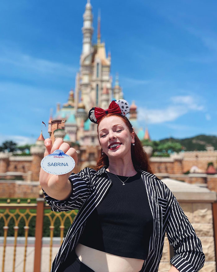 Ex-Disneyland Evil Queen Sabrina smiling in front of castle, holding name tag, sharing truth behind controversial firing.