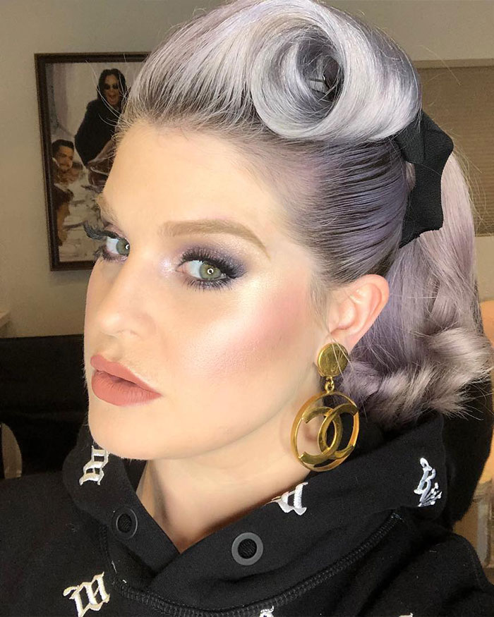 Kelly Osbourne with styled lavender hair, bold makeup, and gold earrings drawing Hollywood attention in a close-up portrait.