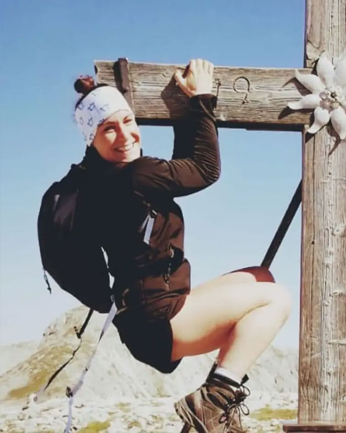 Climber hanging from wooden cross on mountain, smiling and wearing hiking gear and headband under clear blue sky.
