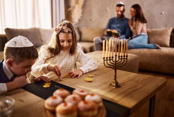 Children playing with a dreidel during Hanukkah, with a menorah and festive treats on the table in a cozy living room. Children playing with a dreidel during Hanukkah, with a menorah and festive treats on the table in a cozy living room.