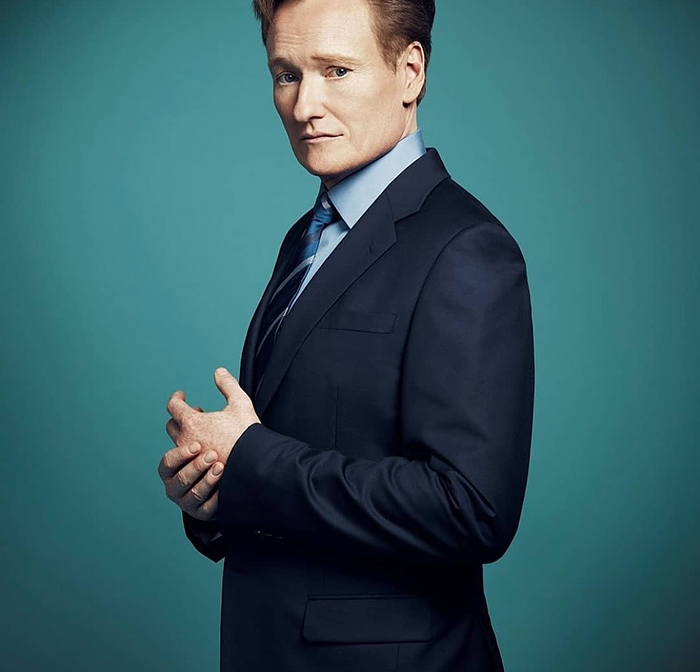 Conan O&rsquo;Brien in a dark suit posing against a teal background ahead of hosting Oscars 2026 event announcement.