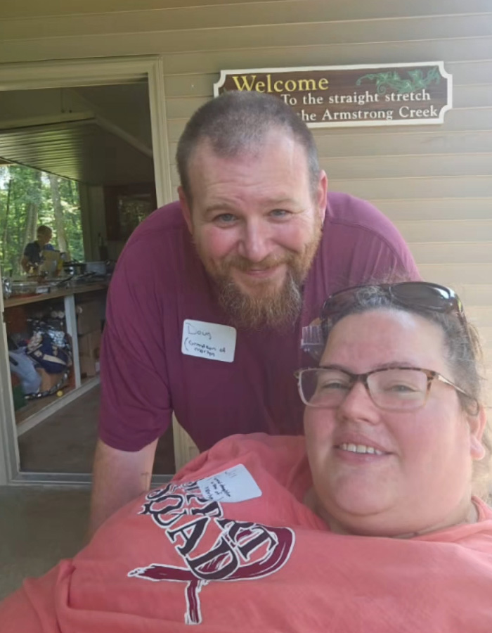 Heartbroken mom with glasses and man with beard smiling indoors near a sign, relating to adoptive father slaying case.