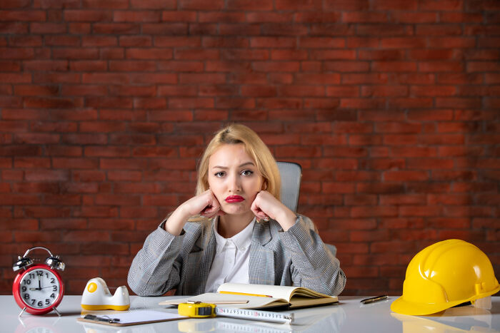 Young woman at desk with alarm clock and hard hat, showing frustration as a brutal wake-up call moment.