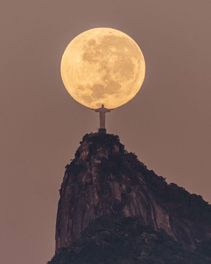 Full moon perfectly aligned behind Christ the Redeemer statue on a mountain, showcasing unreal and incredible reality.