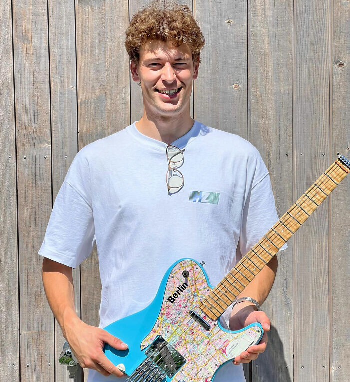 Young man holding a blue electric guitar with a Berlin map design, smiling against a wooden backdrop.
