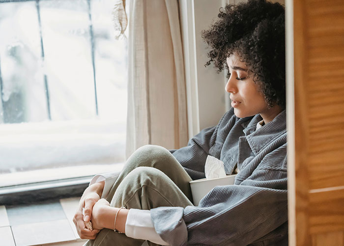 Young woman sitting by window, appearing thoughtful and sad, reflecting on family and relationship challenges.