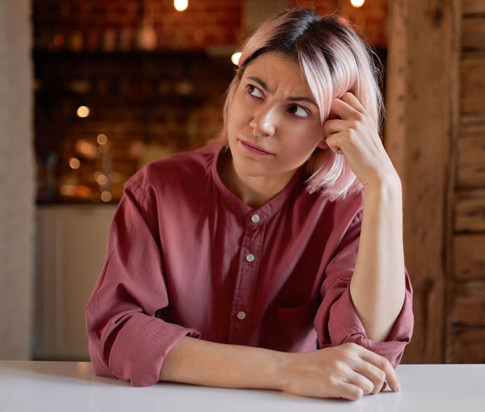 Young woman with pink hair in casual shirt looking thoughtful indoors, reflecting on spicy books causing a stir at a party.
