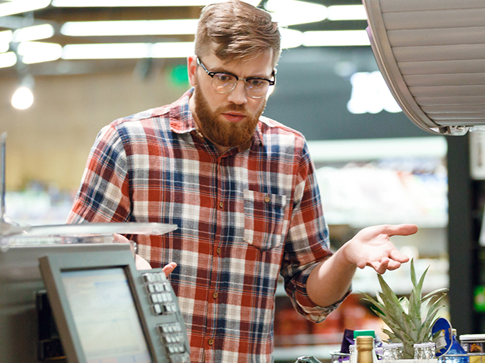 Cashier in a plaid shirt looking confused while working at a grocery store checkout during a crisis situation.