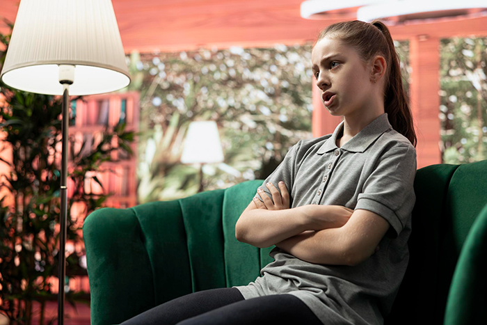 Sassy teen girl sitting on green couch with arms crossed, expressing attitude while talking in a bright living room setting.