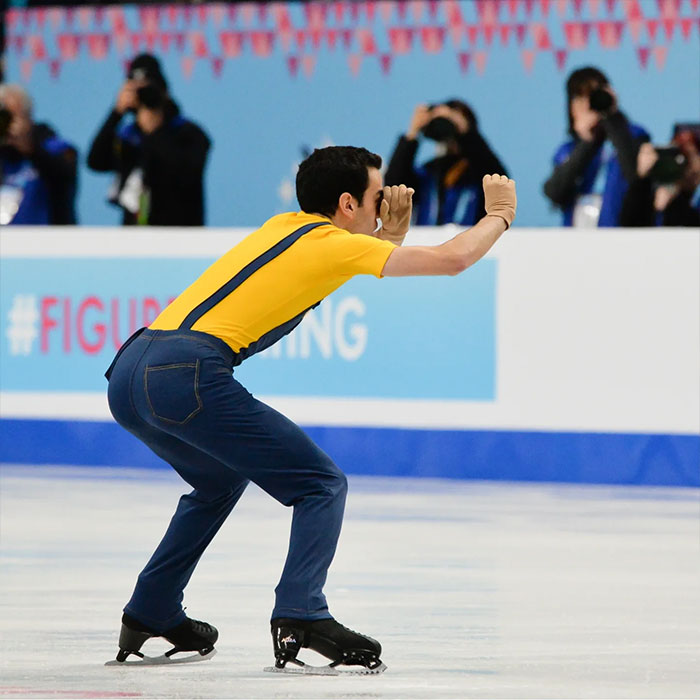 Male Olympic figure skater in a yellow shirt and jeans performing a unique routine on the ice during Winter Games competition.