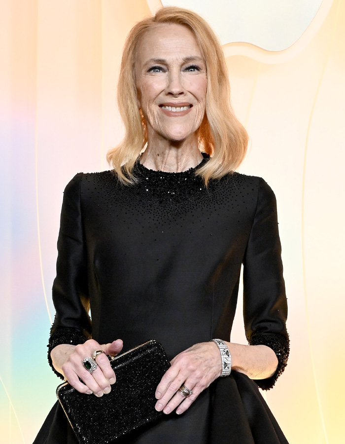 Catherine O'Hara wearing a black dress and holding a black clutch, smiling at a formal event with a light background.