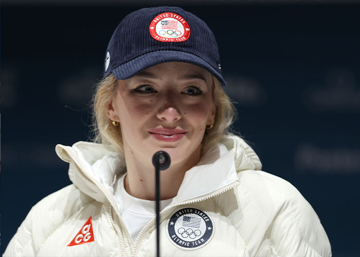 Amber Glenn speaking at a press conference, wearing a US Olympic Team jacket and cap, addressing backlash and threats.