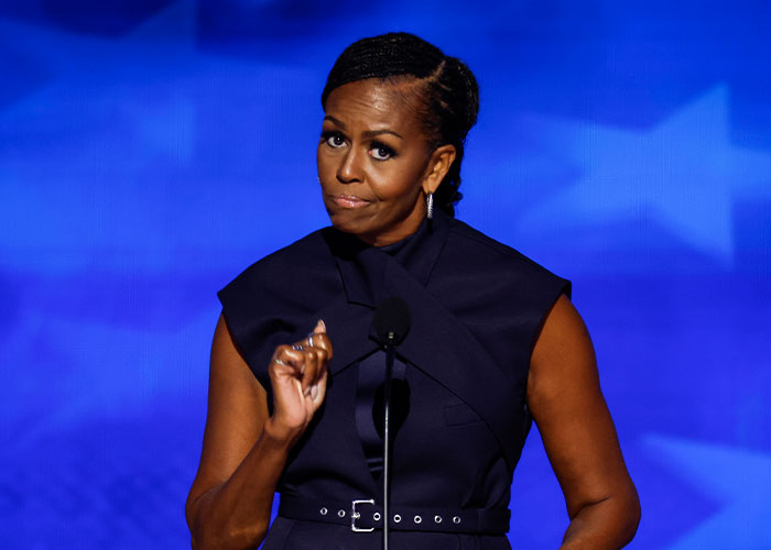 Michelle Obama speaking at a podium during a public event with a blue star-patterned background.