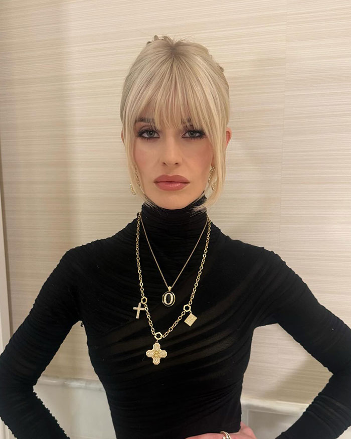 Kelly Osbourne posing in a black top with layered gold necklaces and styled blonde hair in an indoor setting.
