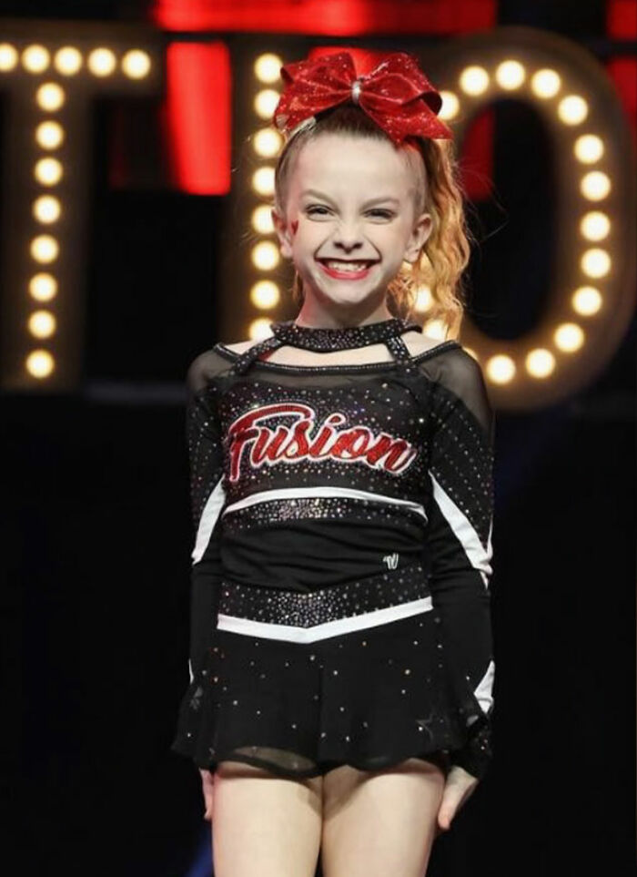 Young cheerleader in a Fusion uniform smiling on stage, related to Utah dance mom&rsquo;s mean texts and parent clash.