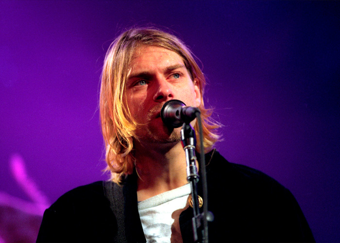 Kurt Cobain singing on stage with a microphone, spotlight highlighting his face against a purple background.