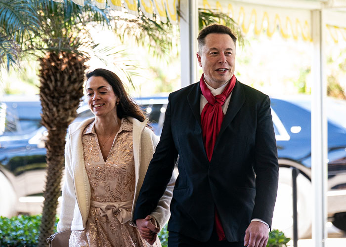 Elon Musk walking outdoors holding hands with a woman, dressed in a black suit and red scarf, sparking bad theories.