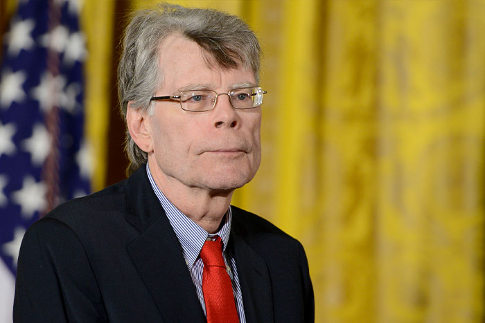 Stephen King in a suit with red tie standing before a blurred American flag and yellow curtain backdrop.