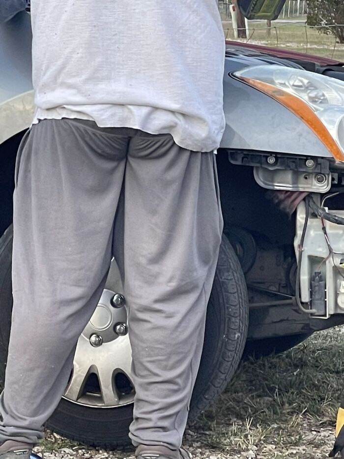 Man standing awkwardly in loose pants near a car with exposed front bumper showing awkward body language.