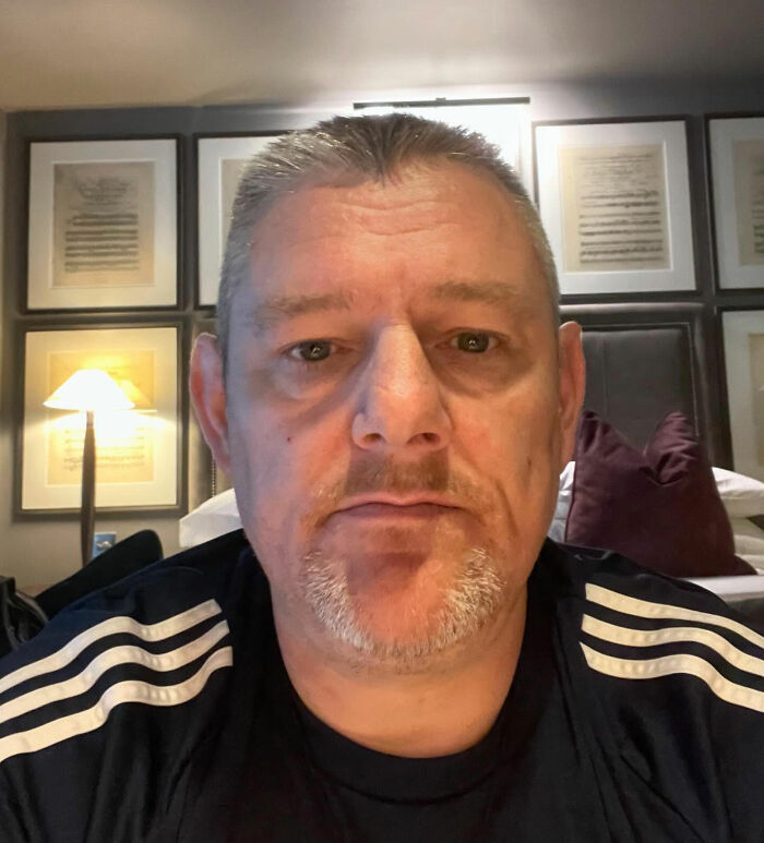 Middle-aged man in a black and white shirt sitting indoors with wall art and a lamp behind him, no visible reaction to incident.