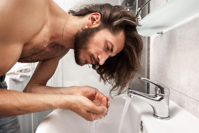 Man with long hair washing face at bathroom sink, reflecting on wild dating experiences that made him question reality.