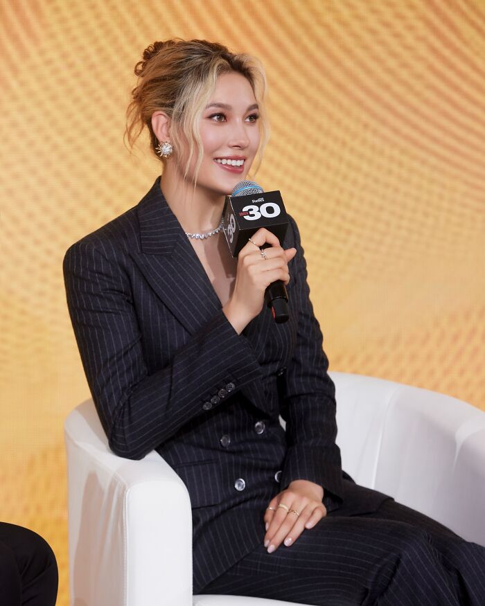 Eileen Gu speaking at an event, dressed in a pinstripe suit, holding a microphone during a panel discussion.