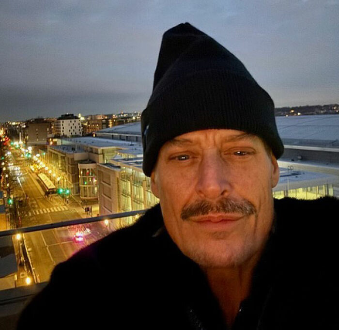 Kid Rock wearing a black beanie and coat, taking a selfie on a rooftop overlooking a city street at dusk.