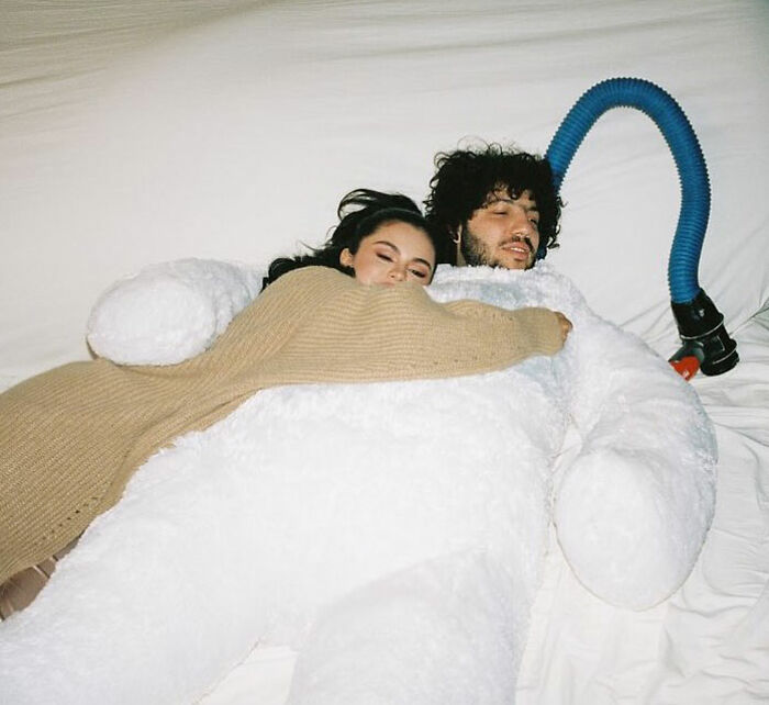 Selena Gomez lying on a bed hugging a man in a large white costume, related to Benny Blanco viral interview topic.