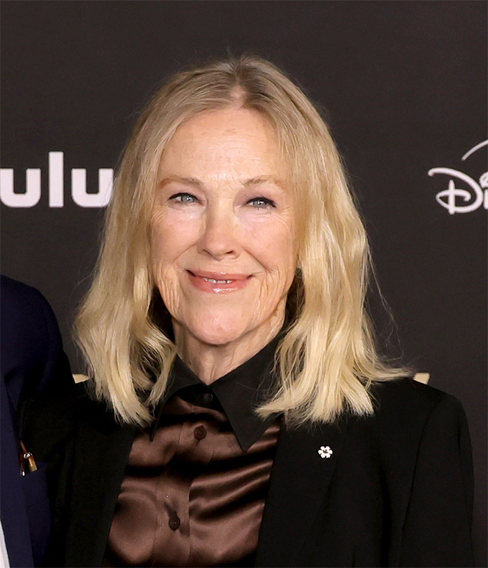 Catherine O'Hara smiling at an event, wearing a black blazer and brown blouse against a dark backdrop.