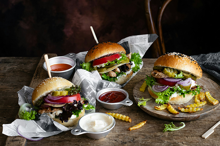 Three gourmet burgers with fries and sauces on a rustic table, highlighting strict diet challenges among friends.