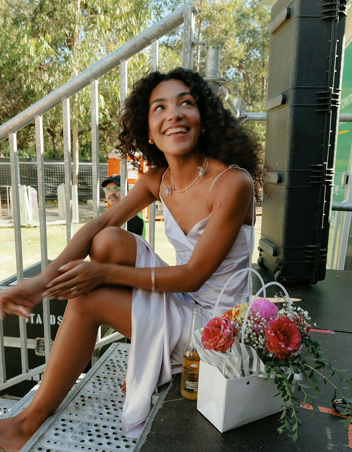 Olivia Dean smiling and sitting outdoors in a white dress with a bouquet of flowers and a drink nearby.