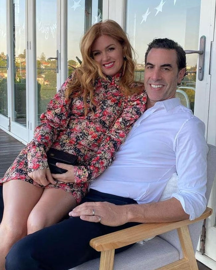 Sacha Baron Cohen and Isla Fisher sitting closely on a chair outdoors, smiling and relaxed together.