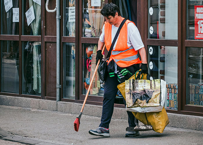 Street cleaner in orange vest sweeping pavement near shop windows showing practical things countries have that make sense.