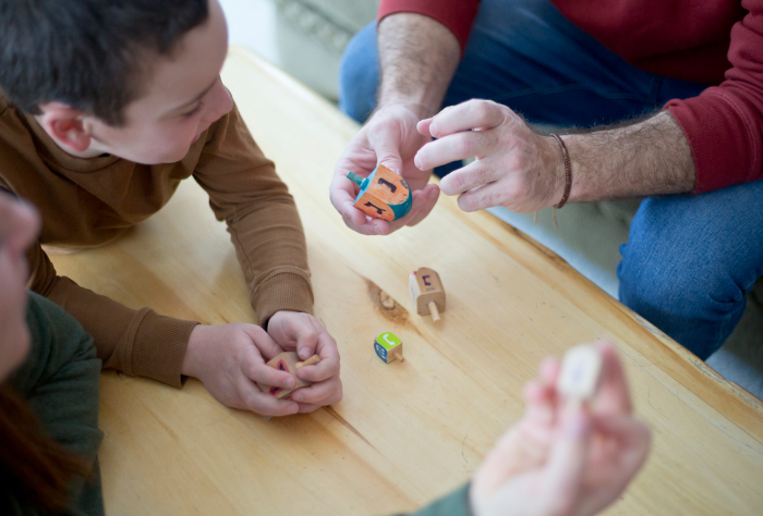 Family playing with a colorful Google dreidel, enjoying a modern Hanukkah activity around a wooden table. Family playing with a colorful Google dreidel, enjoying a modern Hanukkah activity around a wooden table.