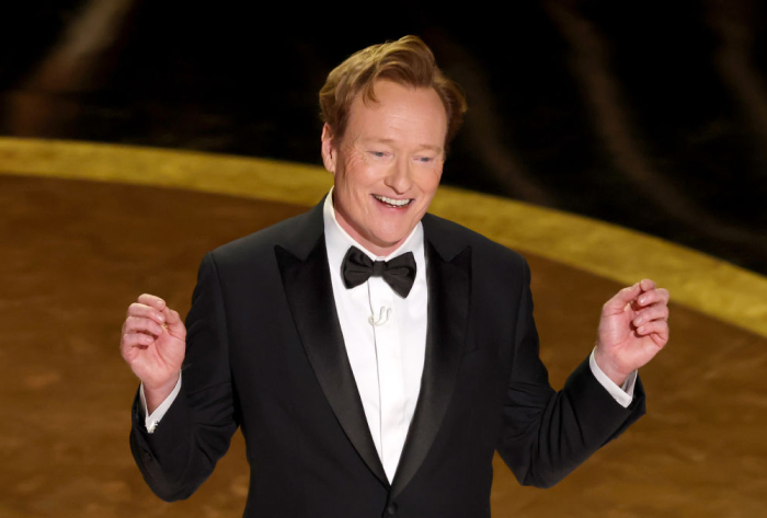 Conan O&rsquo;Brien smiling in a tuxedo on stage, hosting an event related to Oscars 2026 return.