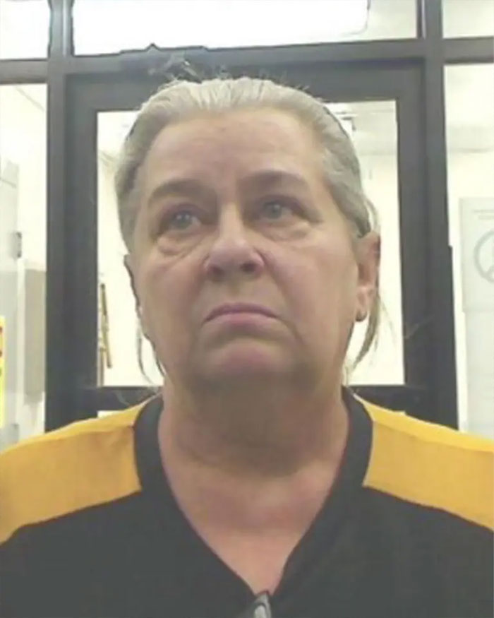 Middle-aged woman with gray hair in a black and yellow shirt, arrested after vanishing 25 years, mugshot in holding area.