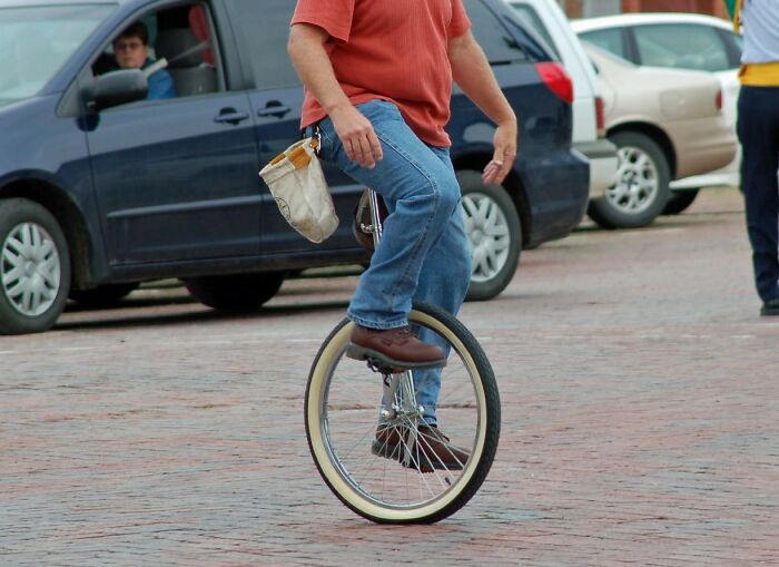 Man riding a unicycle on a brick street, one person watching from a vehicle, capturing a strange witnessed moment.