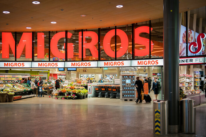 Entrance of a Migros supermarket with shoppers and fresh produce, illustrating everyday facts that come in clutch in conversations