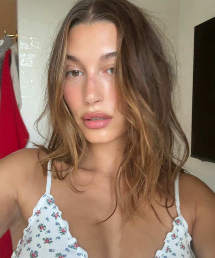 Hailey Bieber wearing lingerie-style top with floral pattern and natural makeup in a casual indoor setting.