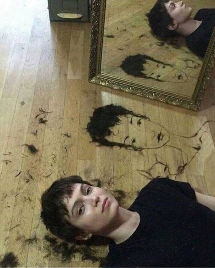 Person lying on wooden floor with hair art drawing matching their haircut next to a mirror, funny weirdly hilarious image.