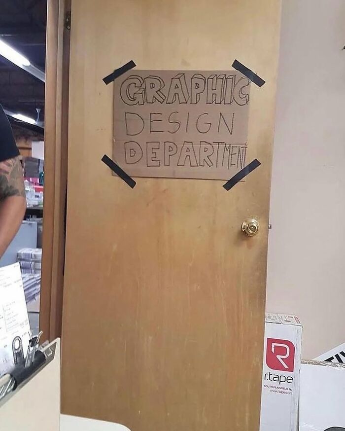 Handwritten cardboard sign taped on wooden door reads Graphic Design Department in casual workplace, weirdly hilarious image.
